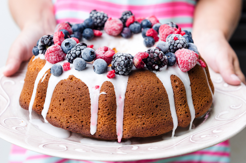 Woman hands holding vanilla bundt cake dessert with icing glaze topped with mixed berries raspberry blueberry blackberry