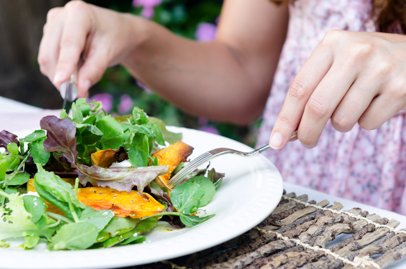 Pair of woman's hands holding cutlery eating a fresh healthy raw spring salad with butternut pumpkin squash at a casual outdoor dining meal location